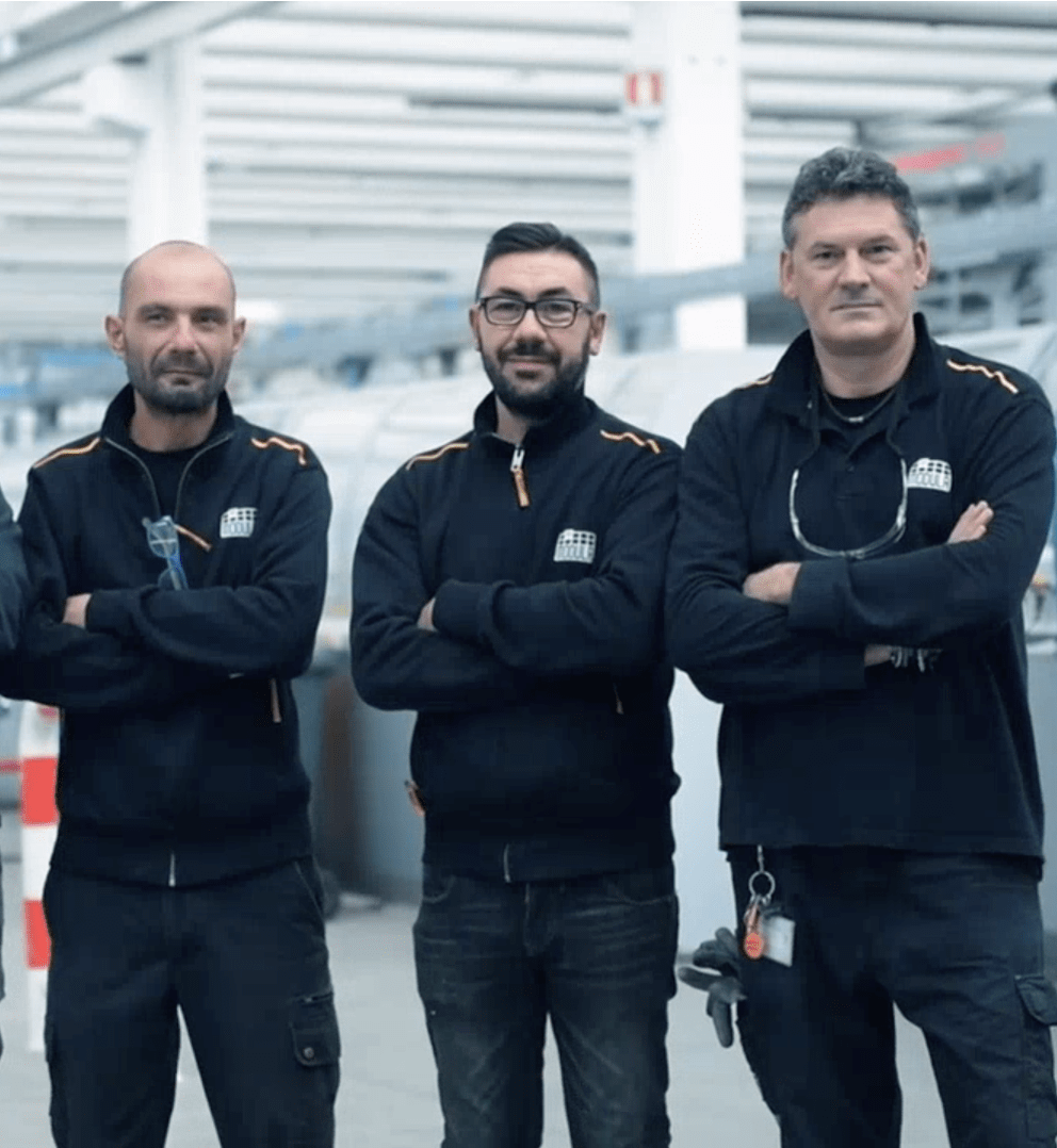 Modula Customer Care team in dark work uniforms stand with arms crossed in a Modula Service factory setting, facing the camera.