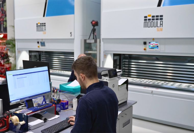New Fer employee managing inventory at a computer workstation with two Modula Slim vertical lift modules in the background.